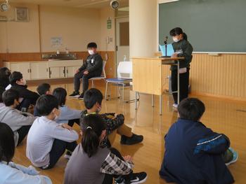 黒板の前のマイク付きの演台に一人の児童が立ち、原稿を見ながら話をしている様子を他の児童たちは床に座って静かに聞いている様子を写した写真