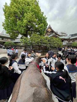 神社と思われる場所で、多くの制服姿の学生たちが銅像を囲んで楽しそうに記念撮影をしている様子の写真