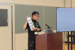 演台に立つ男性教師が紙を手に持ち、プール学習の約束について話している様子の写真