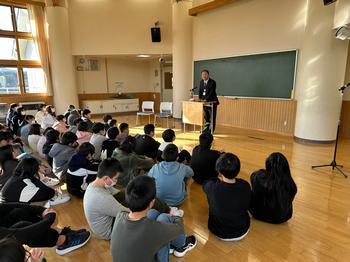 教室の床に座ったたくさんの児童たちが、教卓に立って話す校長先生の話を聞いている様子の写真