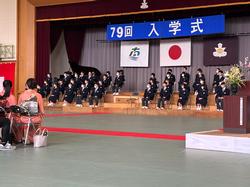 体育館で保護者らが着席する中、舞台には「第79回 入学式」の横断幕が掲げられ、階段状に並んだ椅子に制服姿の多くの生徒が座っている写真