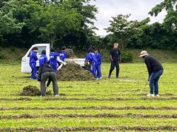 広い芝生の校庭で、ジャージ姿の生徒たちと大人が協力し、軽トラックの荷台に大量の刈り取られた草を積み込んでいる奉仕作業の様子の写真