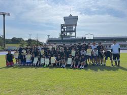 青空が広がる陸上競技場の芝生の上で、黒いユニフォームやTシャツを着用し、賞状を持った生徒を含む大勢の選手の皆さんが集まって写っている集合写真