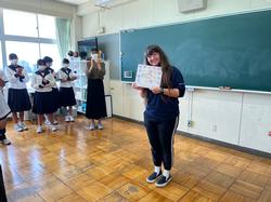 ケイティ先生がメッセージの書かれた色紙を持って笑顔で立っており、その後方には制服姿の生徒たちが拍手をして見守っている写真