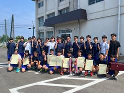 屋外の校舎の前で、体育着姿の男子生徒と女子生徒が、賞状やトロフィー、優勝旗などを持って記念撮影をしている写真