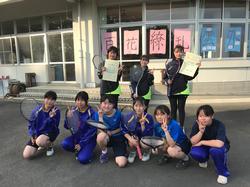 「百花繚乱」の文字が書かれた紙が貼られた窓の前に、ジャージを着た女子生徒たちがラケットを持って並び、後列の2人が表彰状を掲げて写した記念写真