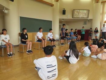 全校児童が座って見守る中、選手の児童が前方に並んだ椅子に座り、中央の児童が自席に立って話をしている水泳記録会壮行式の様子を写した写真