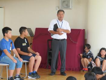 白いポロシャツを着た先生が椅子に座っている選手の児童と選手の前に座っている全校児童に向けて話をしている様子の写真