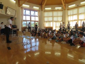 全校生徒が床に並んで座っており、その前に立った校長先生が話をしている様子を写した写真