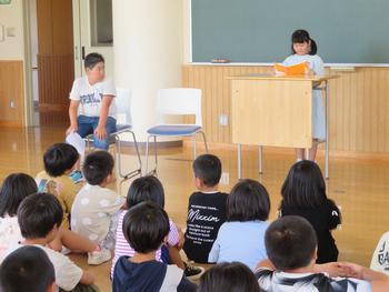 一人の女子児童が教卓の前に立ってオレンジ色の本を読み上げて発表しており、全校生徒が床に並んで座り、その話を静かに聞いている様子を写した写真