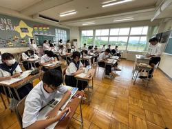 夏服の学生たちが学校の教室で、机に向かい一斉にテストや自習に取り組んでおり、教室内には数名の保護者が立って見守る様子の写真