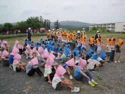 2次避難で、子どもたちがピンク・青・オレンジ色の防災頭巾をかぶり、2列に並んでしゃがんでいて、次々と生徒たちが避難場所に集まっている避難訓練の様子の写真