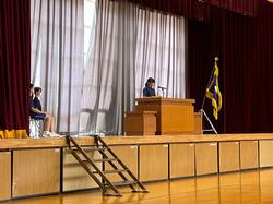 ステージの左側に数名の生徒が座り、中央の演台に一人の女子生徒がt立っている写真