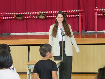 新しいALTの先生がマイクの前に立ち、全校児童に挨拶をしている様子の写真