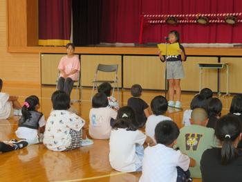 体育館のステージ前で、代表の2人の児童のうち女子児童がマイクの前に立ち、2学期の抱負を話し、全校児童が座って話を聞いている始業式の様子の写真