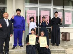 「百花繚乱」の文字が書かれた紙が貼られた窓の前に、黒い袴や学校のジャージを着た生徒たちとスーツの男性が並び、前列の女子生徒2人が表彰状を掲げて写した記念写真