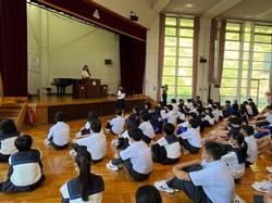 集会場に集まった生徒たちが整列して床に座り、前方のステージ前に立った女子生徒とステージの演台に立った女性の方を見ている様子を後方から撮影した写真