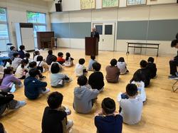 広い部屋で生徒たちが床に座り、演台に立つ大人の方を向いて話を聞いている開校記念集会の様子の写真