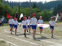 白いTシャツと青い短パンの体操服を着て、赤い帽子をかぶって先頭の児童が日の丸を掲げその後ろに数名の児童が白旗や赤旗を持って、整列して元気に運動場のトラックを行進する小学生たちの写真