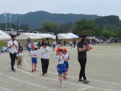 赤い帽子をかぶった児童と保護者方々がグラウンドで大きな円を作り、入谷小唄を踊っている様子の写真