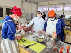 エプロン、三角巾、マスクを身に着けた2人の生徒が、白い三角巾とマスク、白衣を身に着けた女性の指導の下、材料を切ったり調理実習をしている写真