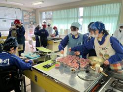 エプロン、三角巾、マスクを身に着けた生徒たちが、調理台にたくさん並べたピンク色のおかずカップに作った料理を盛り付けている写真