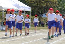 児童たちが白い体操服と青い短パン、赤い帽子を着用し全員が腕を大きく振って一糸乱れず歩いており、規律正しい行進をしている行進の写真