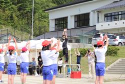 赤い帽子をかぶった児童たちが教師の指導のもと、手を挙げて準備体操をしている様子の写真