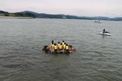 黄色い救命胴衣を着た子どもたちが手作りいかだに乗って水上を進み、活動を行っている写真