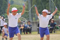 白と赤の帽子をかぶった2人の男子児童が並んで縄跳びをし、両腕を高く挙げてジャンプする瞬間が写っている写真