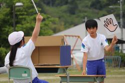 白い帽子をかぶった女子児童が運動場に設置された机の前に座り、棒を持った手を高く挙げており、立っている男子児童が片手に大きな手のイラストを掲げてやりとりしている様子の写真