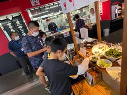 児童たちがビュッフェ形式のカウンターに並べられたいくつかの大きなボウルに入った野菜や薬味をトングを使い料理を皿に取り分けており、その後ろに大人の男性が並んでいる写真