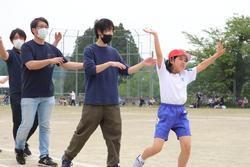 一番前の女子児童は赤い帽子をかぶり両手を大きく上げて、笑顔で元気よく歩き、大人と児童たちが列になって踊りを踊っている写真