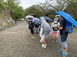「雨の中、児童たちがレインコートや傘をさして鶴ヶ城の石垣の前で見学をしている様子の写真