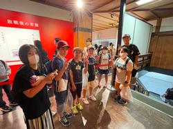 鶴ヶ城の屋内展示室で、赤い壁に展示された資料の前に、児童たちが並んで立ち、カメラに向かってピースサインをしている写真