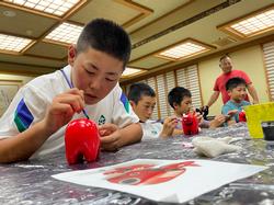 テーブルの上には絵の具の容器や筆、布などが置かれており、集中した様子で一つひとつ丁寧に赤べこに色を塗っている児童たちの写真