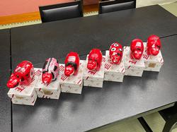 黒いテーブルの上に、赤い張り子の赤べこが7体並べられ、それぞれ紙箱の上に1体ずつ乗せられている写真
