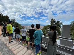 飯盛山から眼下に街並みが広がっており、遠くの景色を見渡している児童たちの後ろ姿だけが写っている写真