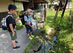 古民家風の建物の側で、3人の児童たちが小さな小川をのぞき込んでいる写真