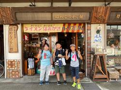 「なつかし屋」という名前の昔懐かしい雰囲気の店の前で、3人の児童が紙袋を持って並んで立っている写真
