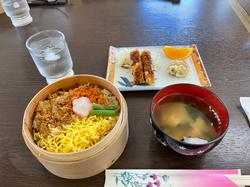 丸い木の器に、色とりどりの食材が盛られたご飯（錦糸卵、野菜、魚そぼろなど）が入っていて、隣に味噌汁と小皿料理（揚げ物、オレンジ、漬物）、そしてお冷が並んだ和食の定食を撮影した写真