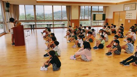演台に立つ代表児童の発表を体育座りをして聞いている児童達の写真