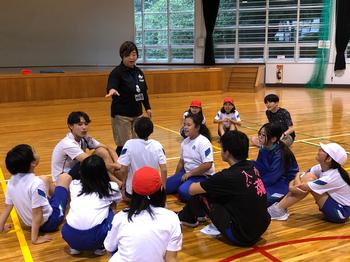 体育館内で先生の説明を座りながら聞いている生徒たちの写真