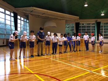 先生と一緒に十数名の生徒が長い棒を床にまっすぐにおいて1列に並んでいる写真