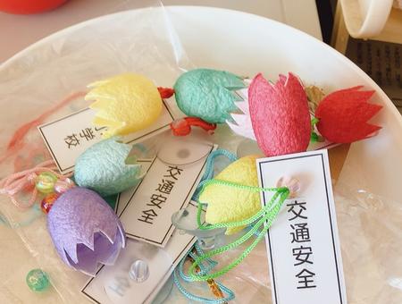 紐の色も様々で、それぞれ「交通安全」と書かれた札が付いている、赤や黄色などのカラフルな色で作られたチューリップの花型の繭マスコットのお守りが、白い皿の上に並べられている写真