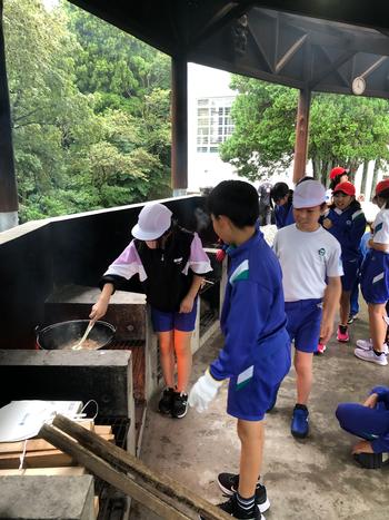 屋外で大きな鍋の料理をかき混ぜている女の子とその様子を見ている男の子たちの写真