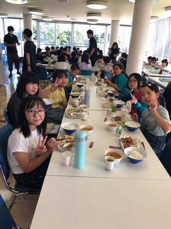 長いテーブルの両側に女子児童が座っており、カメラに向かって笑顔でピースサインをしている食堂で食事をしている様子を写した写真