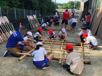 白や赤の帽子をかぶり、体操服を着用した10人以上の小学生たちが、竹の棒を組み合わせて筏のようなものを作っている様子を写した写真
