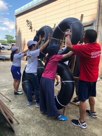 4人の子どもと1人の赤いTシャツを着た男性が大きな黒い浮き輪のようなチューブを竹の骨組みにしっかりとくくりつけ全員で支えている様子の写真