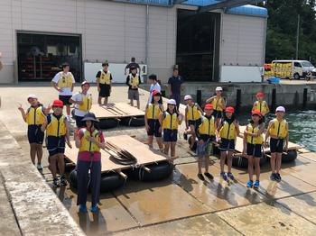 黄色いライフジャケットを着用した20人ほどの小学生たちが港のような場所で筏を前にして並んでいる様子を写した写真